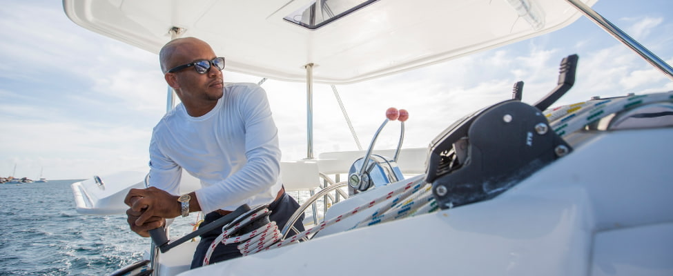 An experienced skipper wearing sunglasses steers a sailing yacht across the open sea. In the background, the deep blue water sparkles under the sun, while the skipper’s focused gaze is directed straight ahead - a symbol of safety, experience, and relaxed charter trips with professional guidance.