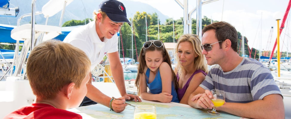 A family sits relaxed around a table with drinks in hand while a captain explains the planned sailing route on an unfolded nautical chart. In the background, a harbor filled with boats sets the scene and conveys the atmosphere of an upcoming charter trip. The image represents shared trip planning, anticipation, and a sailing holiday with a skipper.