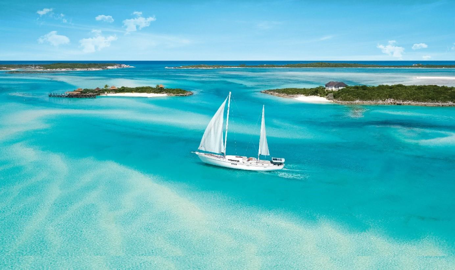 Sailboat cruising in turquoise Bahamian waters with small lush islands in the background, ideal for a yacht charter adventure.
