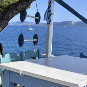 Ein weiß gestrichener Tisch und eine Bank stehen auf einer Terrasse mit Blick auf das Meer. Daneben hängt eine dekorative Leiter mit runden Elementen, und eine griechische Flagge weht sanft im Wind. Im Hintergrund ist das offene Meer zu sehen, auf dem ein Segelboot vorbeifährt, sowie entfernte Inseln oder Küstenlinien. Die Szene strahlt Ruhe, Einfachheit und mediterranes Flair aus.