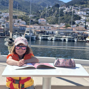 A child sits at a small table on board a boat, drawing or writing in an open notebook. They are wearing a bright orange life jacket, a patterned cap, and sunglasses to shield their eyes from the sun. The child smiles slightly, appearing relaxed and content. On the table beside the notebook is a colourful pencil case. In the background, a picturesque harbour is visible, filled with moored boats and yachts, with clusters of houses climbing up a green hillside. The scene combines a sense of creativity, holiday relaxation, and coastal charm.