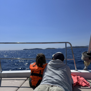 Two people lie side by side on their stomachs on the deck of a boat, looking out over the railing at the deep blue sea. One is wearing an orange life jacket, while the other has on a grey jumper and a cap. The water sparkles in the sunlight, and soft, distant hills or islands stretch along the horizon. The scene conveys a sense of calm curiosity and shared quiet observation.