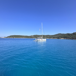 A single sailing boat rests peacefully on clear, turquoise water. The sea is so calm that the boat appears almost reflected on its surface. In the background, a green, hilly coastline extends beneath a cloudless, vivid blue sky. The atmosphere feels tranquil and timeless, far removed from any rush or noise.