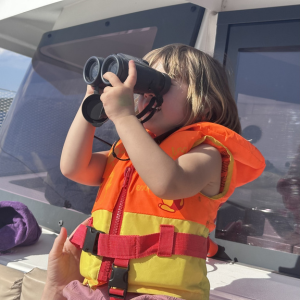 A young child stands on board a boat wearing a bright orange life jacket with yellow and red details. They hold a pair of binoculars up to their eyes with both hands, gazing intently into the distance as if searching for something out at sea. The sunlight highlights their hair, giving it a golden glow. Behind them, parts of the boat’s structure and windows are visible, while an adult’s hand gently supports the child’s arm for balance.