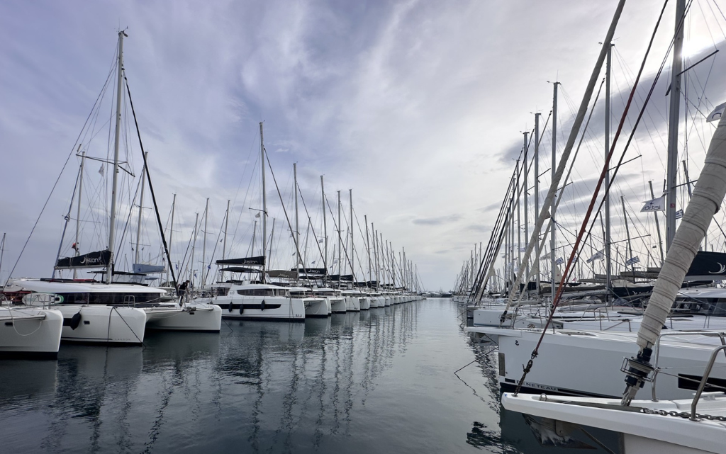 Eine ruhige Marina erstreckt sich unter einem leicht bewölkten Himmel, in der zahlreiche Segelboote dicht nebeneinander an beiden Seiten eines schmalen Wasserkanals festgemacht sind. Die schlanken Masten der Boote ragen in den Himmel und bilden ein dichtes, fast geometrisches Muster aus Linien. Das Wasser ist glatt und spiegelt die Boote sowie die Masten leicht verschwommen wider. Die Szene wirkt still und geordnet, mit einer fast symmetrischen Perspektive, die den Blick in die Tiefe des Hafens lenkt. Es herrscht eine entspannte, beinahe meditative Atmosphäre ohne sichtbare Bewegung.