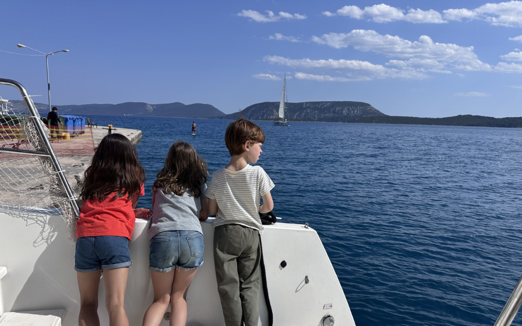 Drei Kinder lehnen sich über die Reling eines Bootes und blicken neugierig auf das Wasser hinaus. Sie tragen sommerliche Kleidung wie T-Shirts und Shorts. Das Meer ist tiefblau und ruhig, während im Hintergrund ein Segelboot vorbeifährt und sich eine hügelige Küstenlandschaft erstreckt. Die Körperhaltung der Kinder zeigt Interesse und Begeisterung, als würden sie etwas im Wasser beobachten oder einfach die Aussicht genießen.
