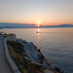 The sun slowly sets over the sea, casting warm orange hues across the sky. A small boat moves across the water in the distance, leaving a delicate trail behind it. In the foreground, a narrow path runs along a rocky coastline with low vegetation and scattered flowers. Silhouettes of mountains or islands can be seen on the horizon, enhancing the peaceful, romantic mood.