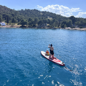 A person stands on a stand-up paddleboard, moving across clear, deep blue water. A child sits in front of them, and both are wearing life jackets. The surrounding area shows a quiet bay with wooded hills and a few buildings along the shore. Sunlight sparkles on the water, creating a sense of leisure, adventure, and shared outdoor activity.